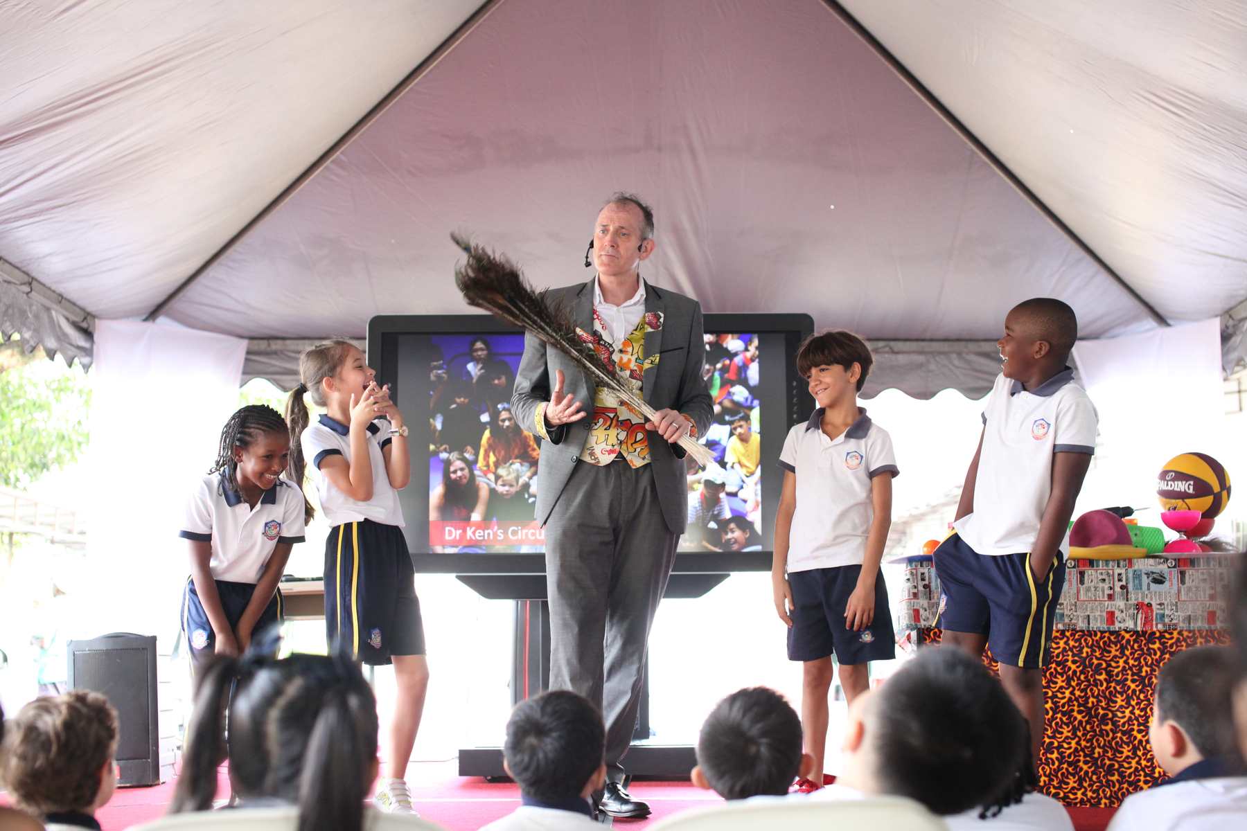 School pupils having fun on stage with Dr Ken Dr Ken’s Circus Science for Schools prepares to explore the science of clowning, holding peacock feathers while his four young volunteers are excited to be on stage.