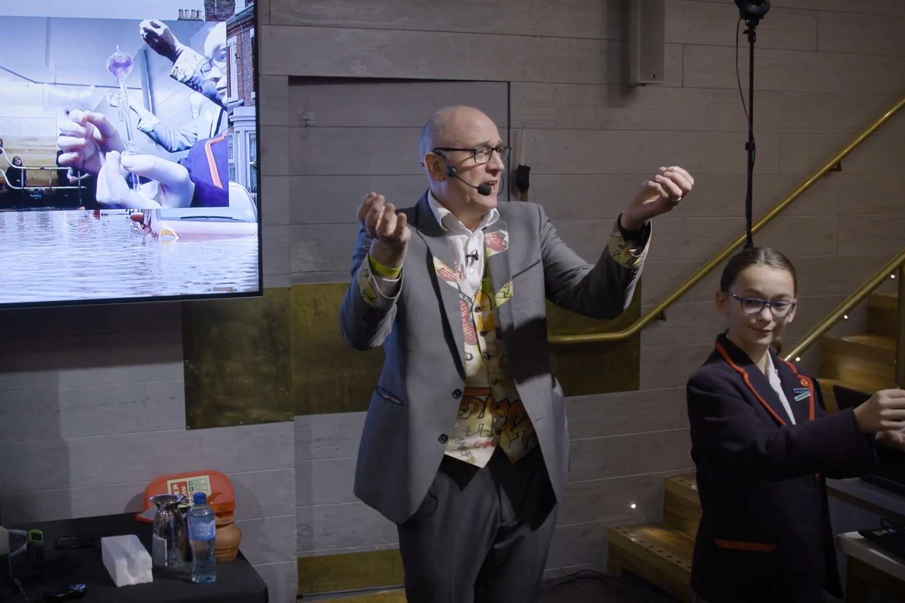 A pupil holds a hand boiler and presents it to a webcam. A screen shows a close up of the volunteers hand and a blue liquid rising up the hand boiler.