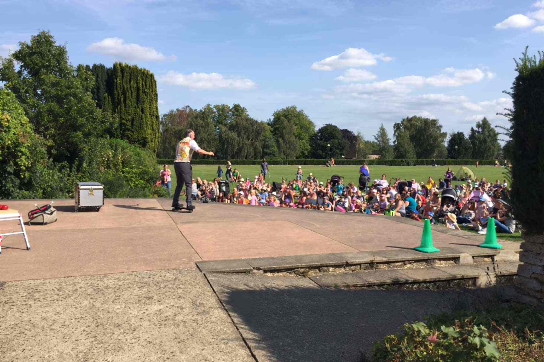 Dr Ken is showing push and pull forces whilst riding snake board in front of an audience sat on the grass in Waterloo park, Norwich.