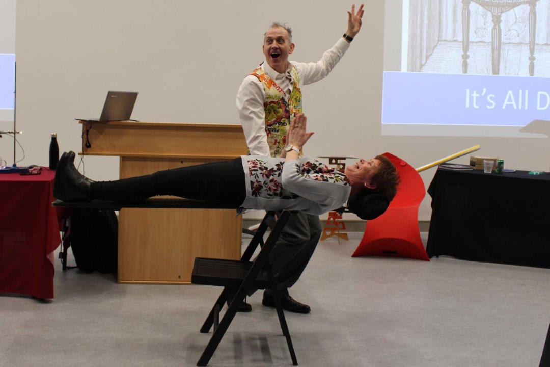 A Teacher lying on a plank of wood is magically balanced on the back of a chair.