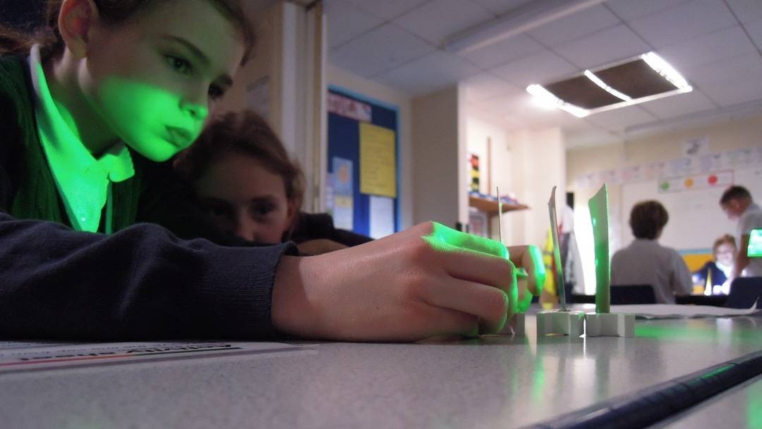 Pupils deep in concentration whilst attempting to see if light travels in a straight line. They shine a green light through 3 vertically aligned identical pieces of card, each card has small holes strategically placed at the same height.